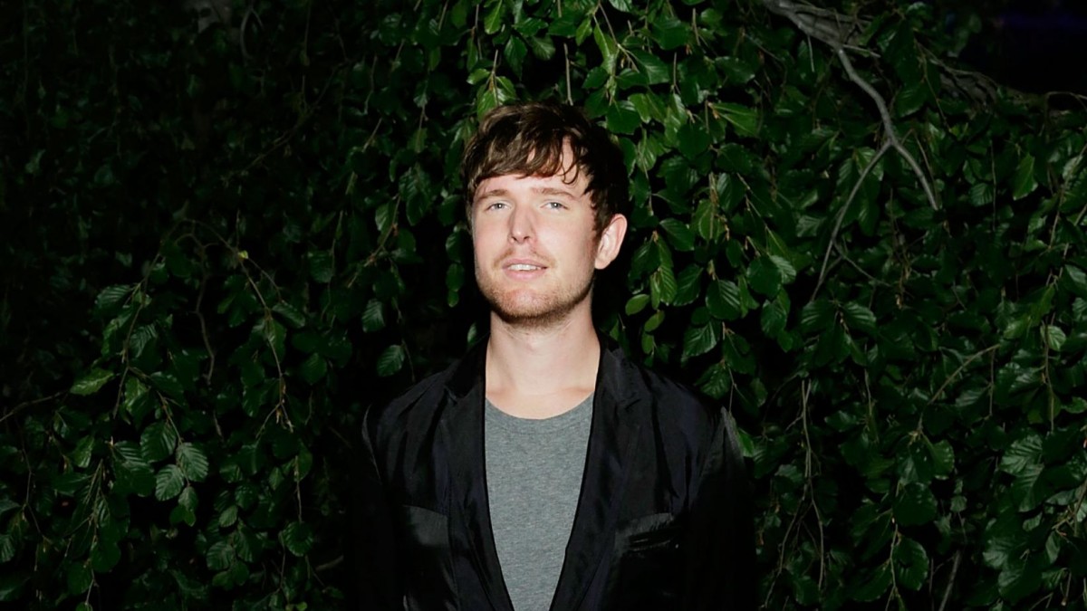 James Blake mengenakan kaus abu-abu, dengan luaran hitam, berdiri di depan pohon.